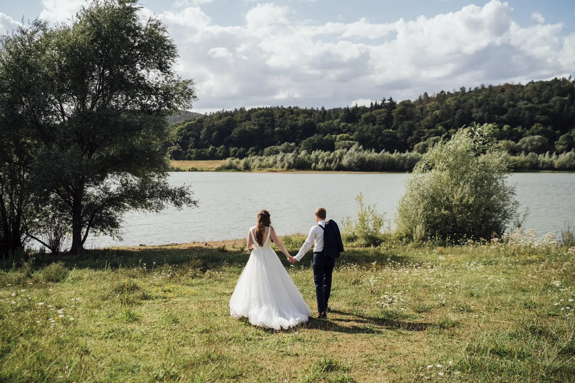 hochzeit-rittergut-meinbrexen-20190810-DSC07057 Brautpaar geht über eine Wiese zum Ufer der Weser hinunter. Der Bräutigam trägt sein Sakko über der Schulter.